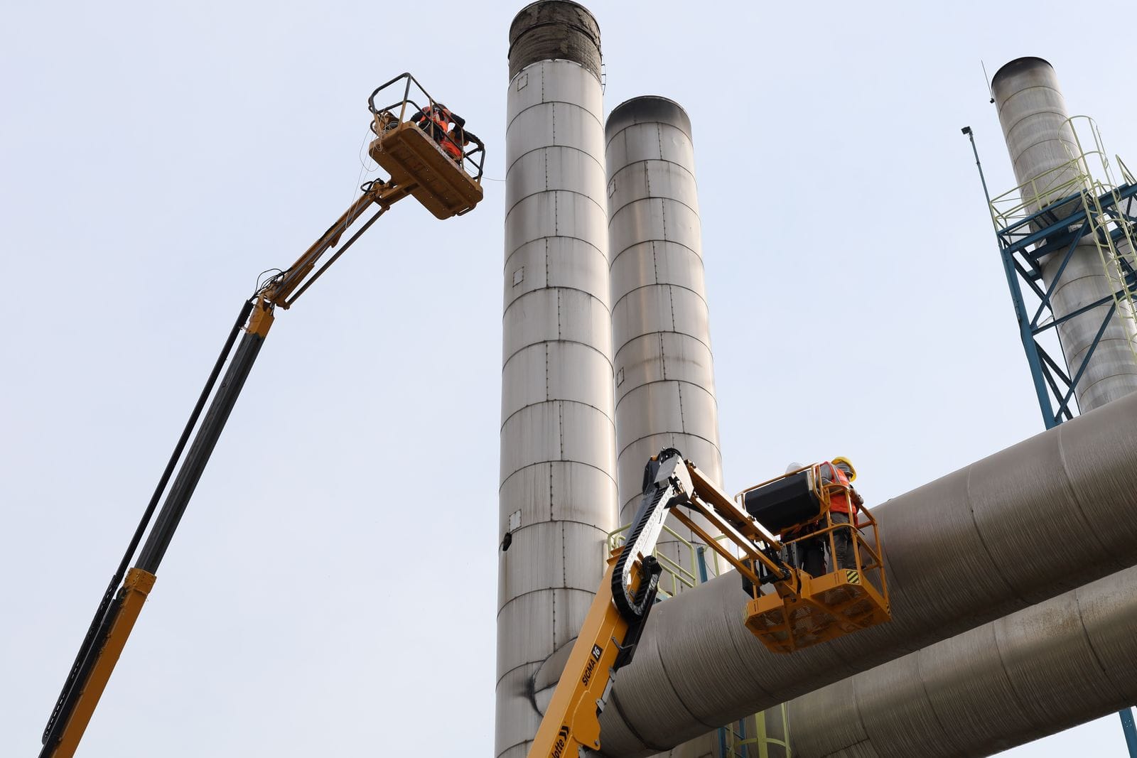 Two aerial work platforms raised beside tall grey chimneys and a large duct during maintenance work at a plant
