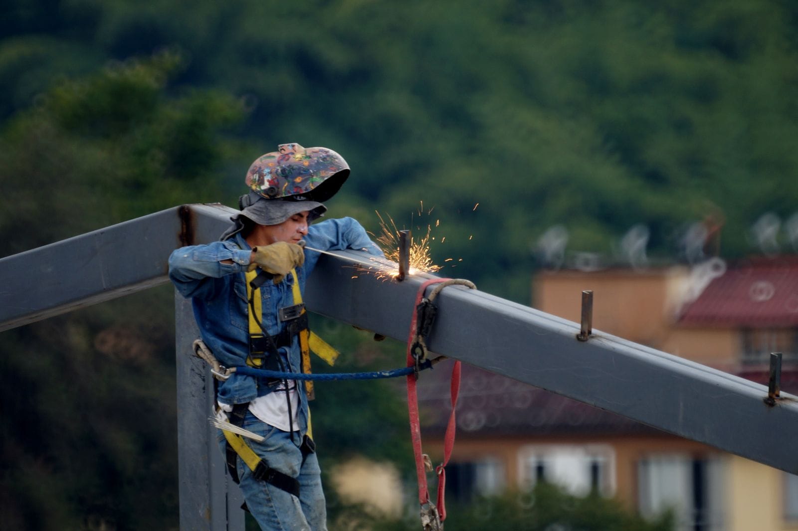 Welder in a protective helmet and safety harness welding a steel beam on site, the joining work used to build a storage tank shell