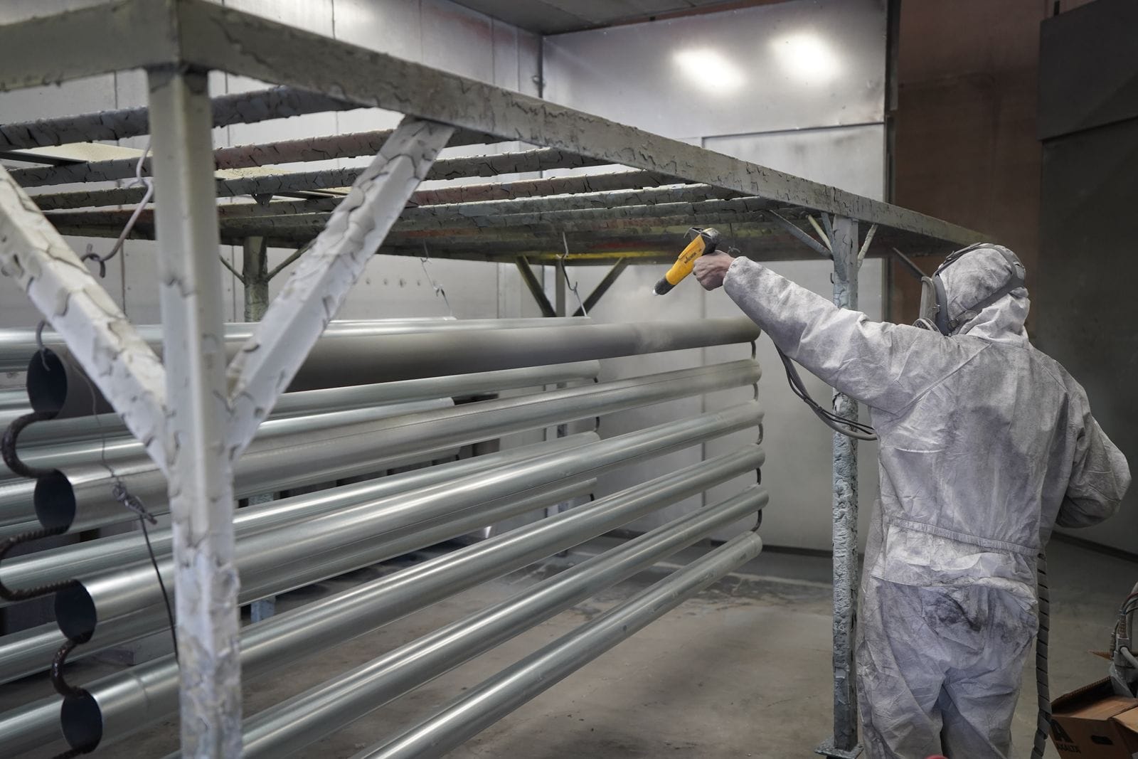Applicator in a full protective suit spraying a coating onto a rack of steel pipes inside a spray booth