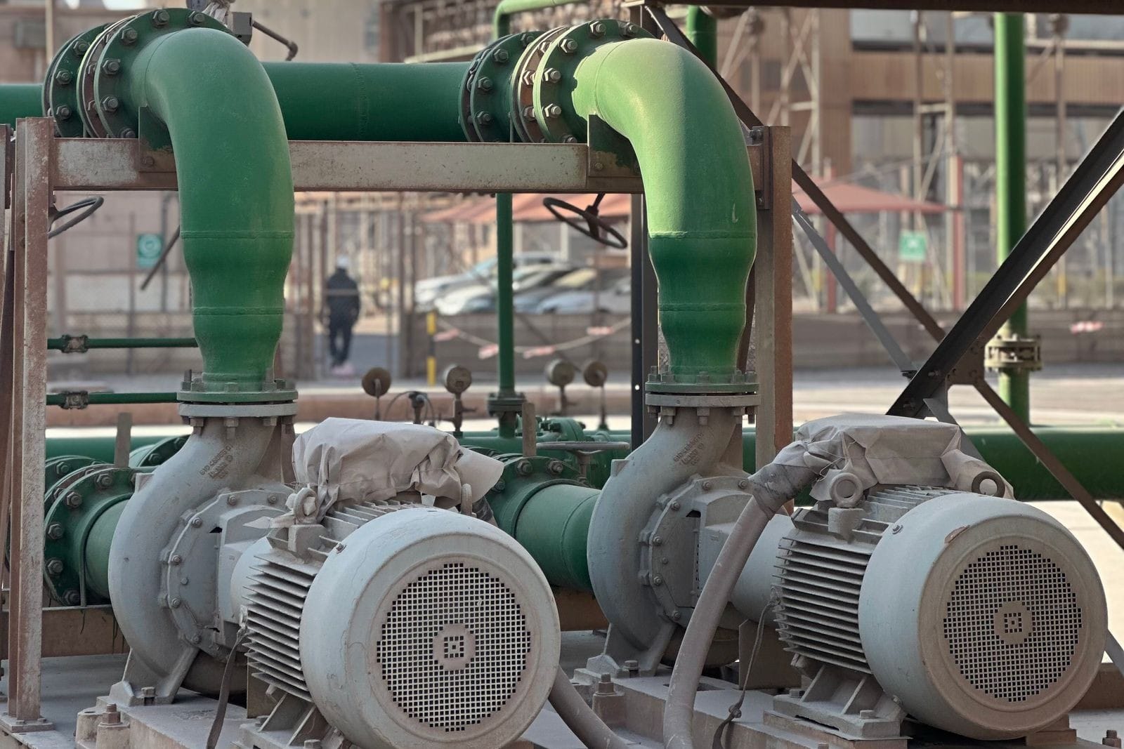 Two centrifugal pumps driven by electric motors with dust covers, connected to large green flanged pipework in an industrial plant