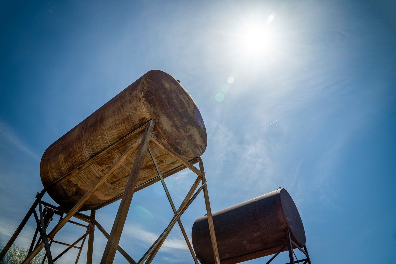 Two rusty steel tanks on tubular steel stands seen from below against a blue sky with the sun overhead, showing heavy surface corrosion