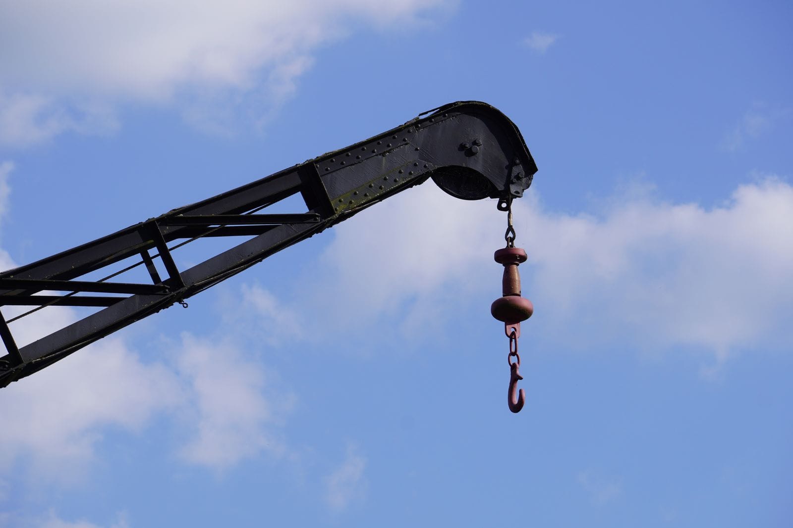 Tip of a lattice crane boom with a hook and ball hanging from it against a blue sky with white clouds