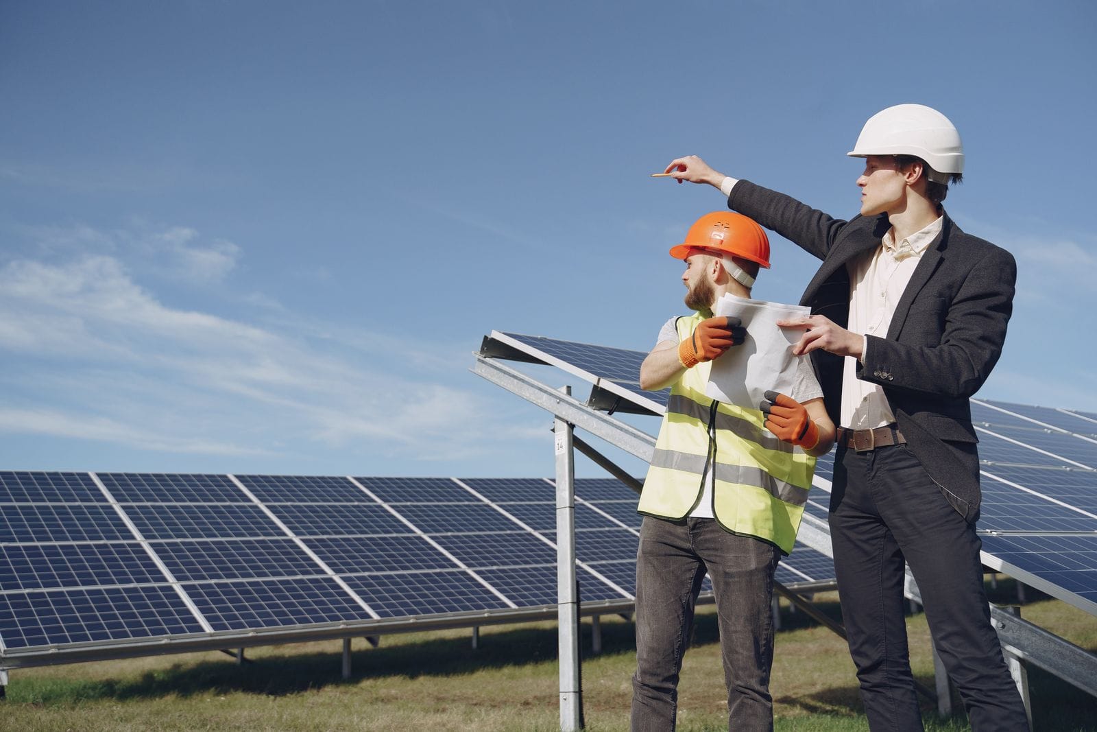 Two people in a hard hat and a safety helmet with a high-visibility vest pointing across a ground-mounted solar array on steel supports