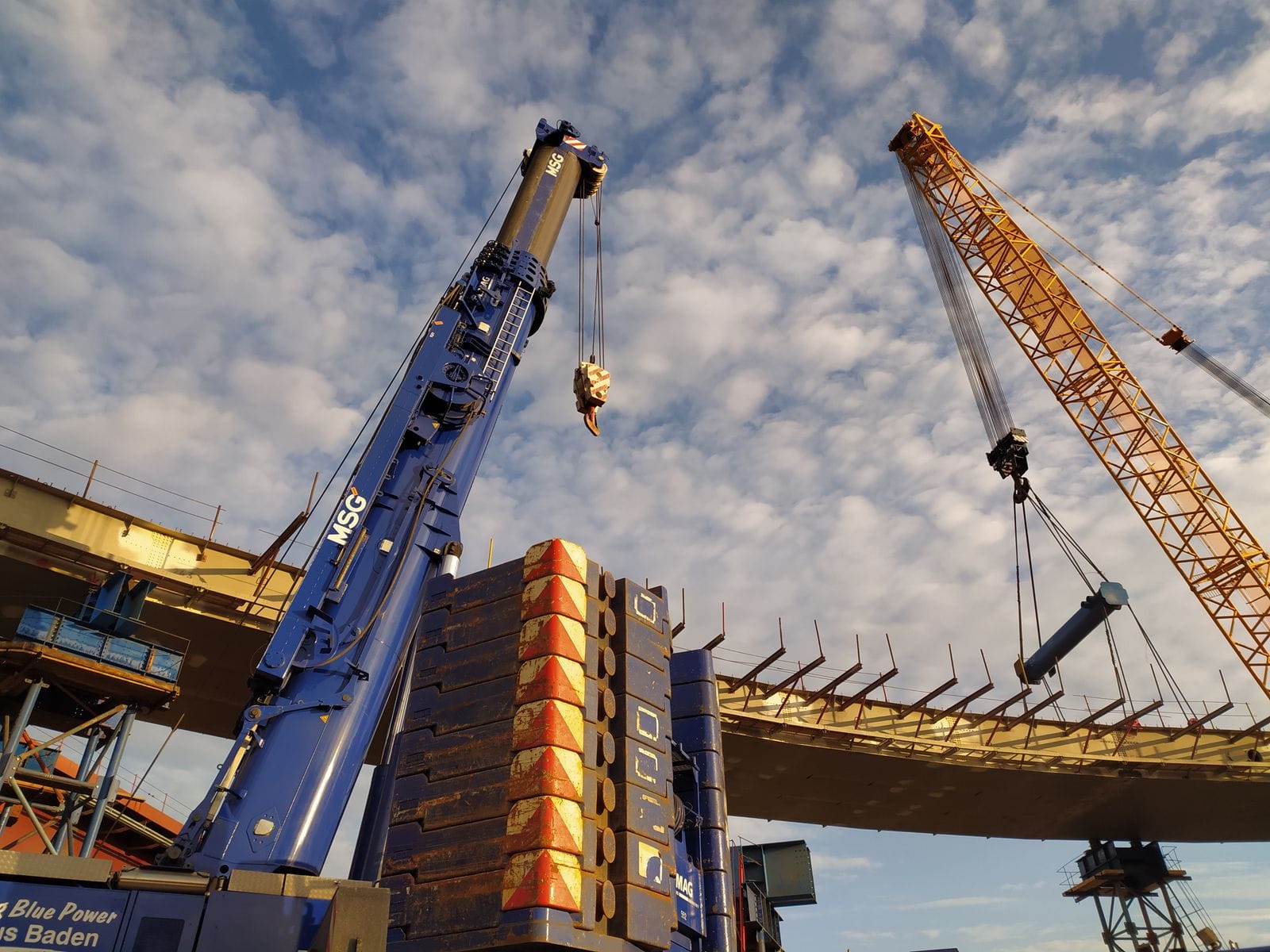 Two heavy cranes lifting steel sections at an industrial construction site, the equipment used to erect tank shell plates