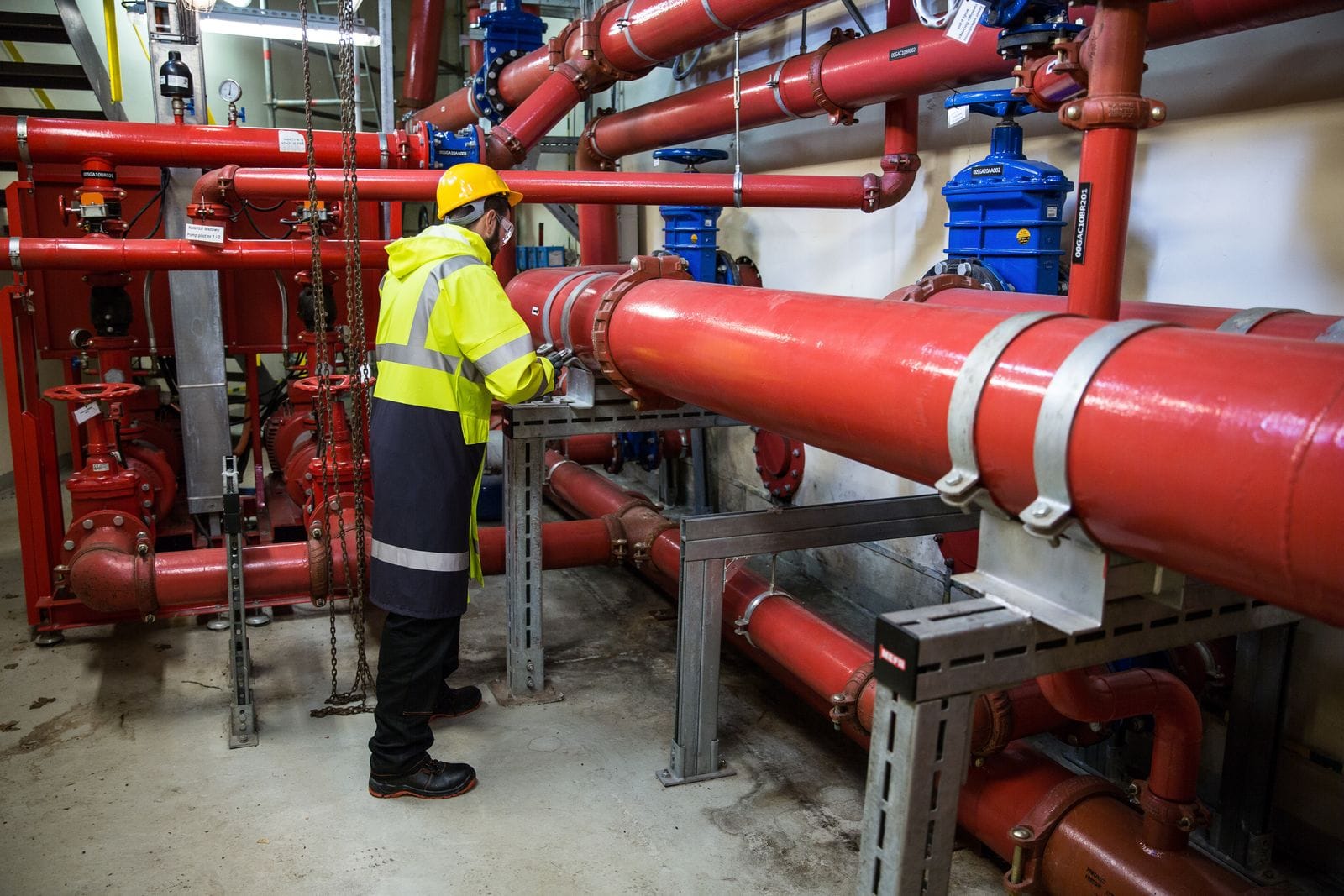 Inspector in a high-visibility jacket and yellow hard hat checking red industrial piping and blue valves in a plant room