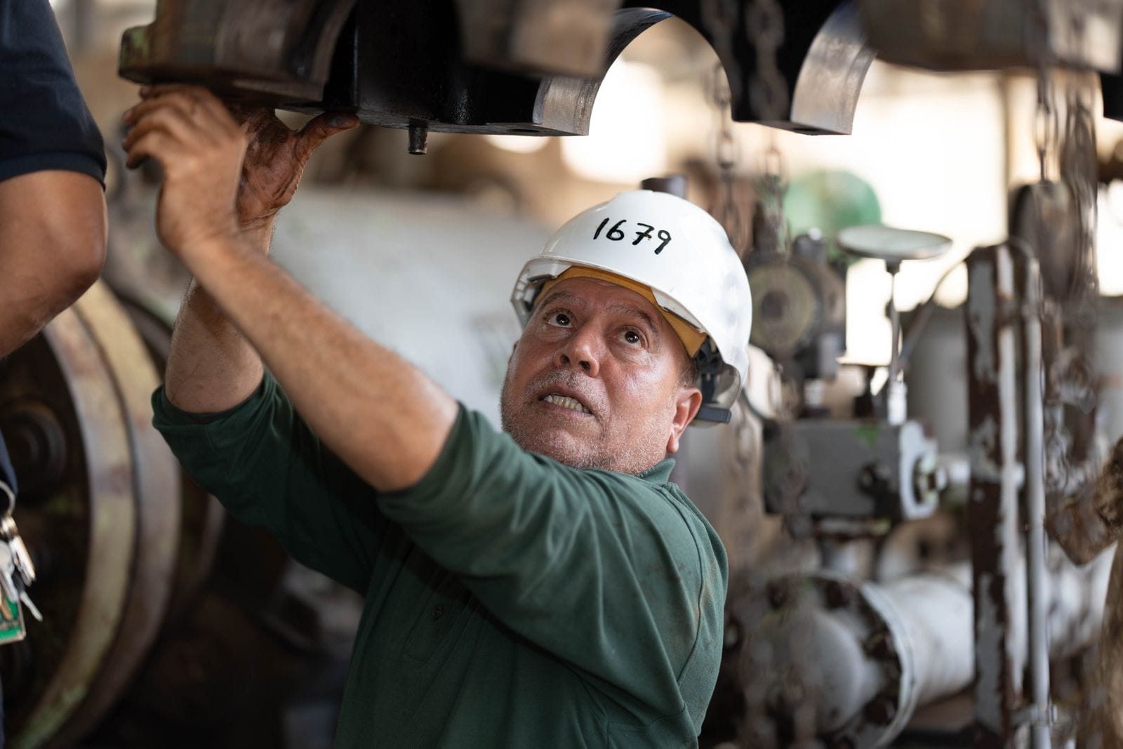 Mechanic in a white hard hat reaching up to inspect heavy machinery components in an industrial plant