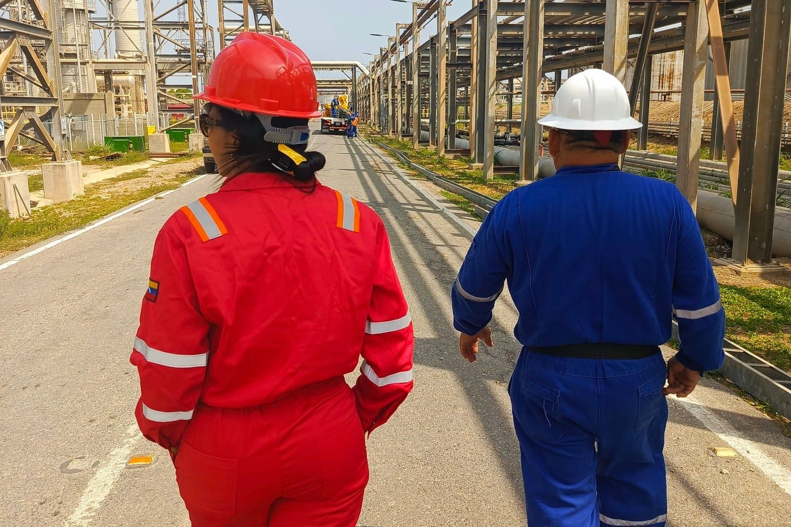 Two workers in red and blue coveralls and hard hats walking along a plant road beside an elevated pipe rack in a refinery