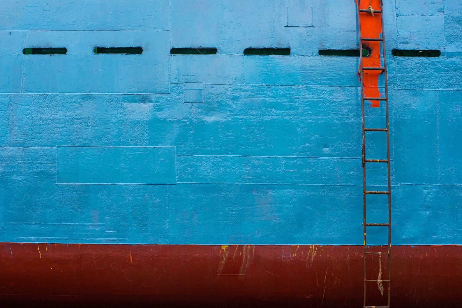 Steel ship hull painted blue above a red waterline band with an orange steel ladder running down the side