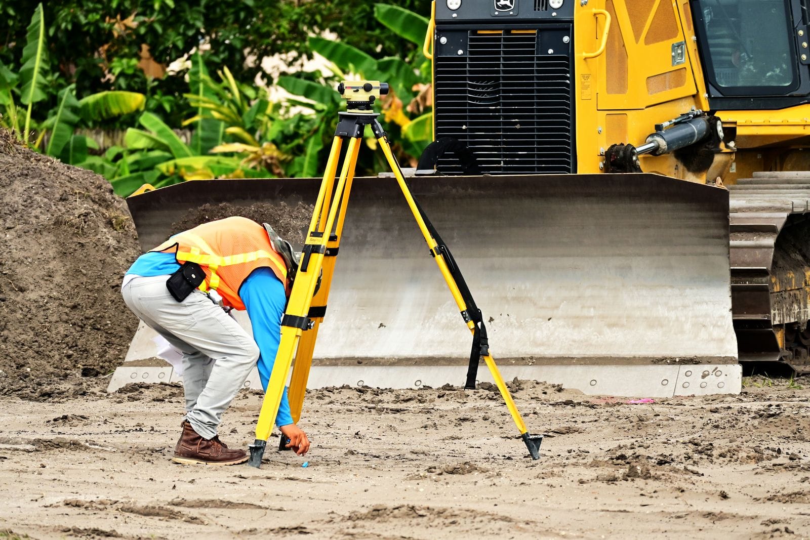 Surveyor setting up a yellow tripod with a level instrument on a sandy construction site with tropical vegetation, used to measure tank settlement