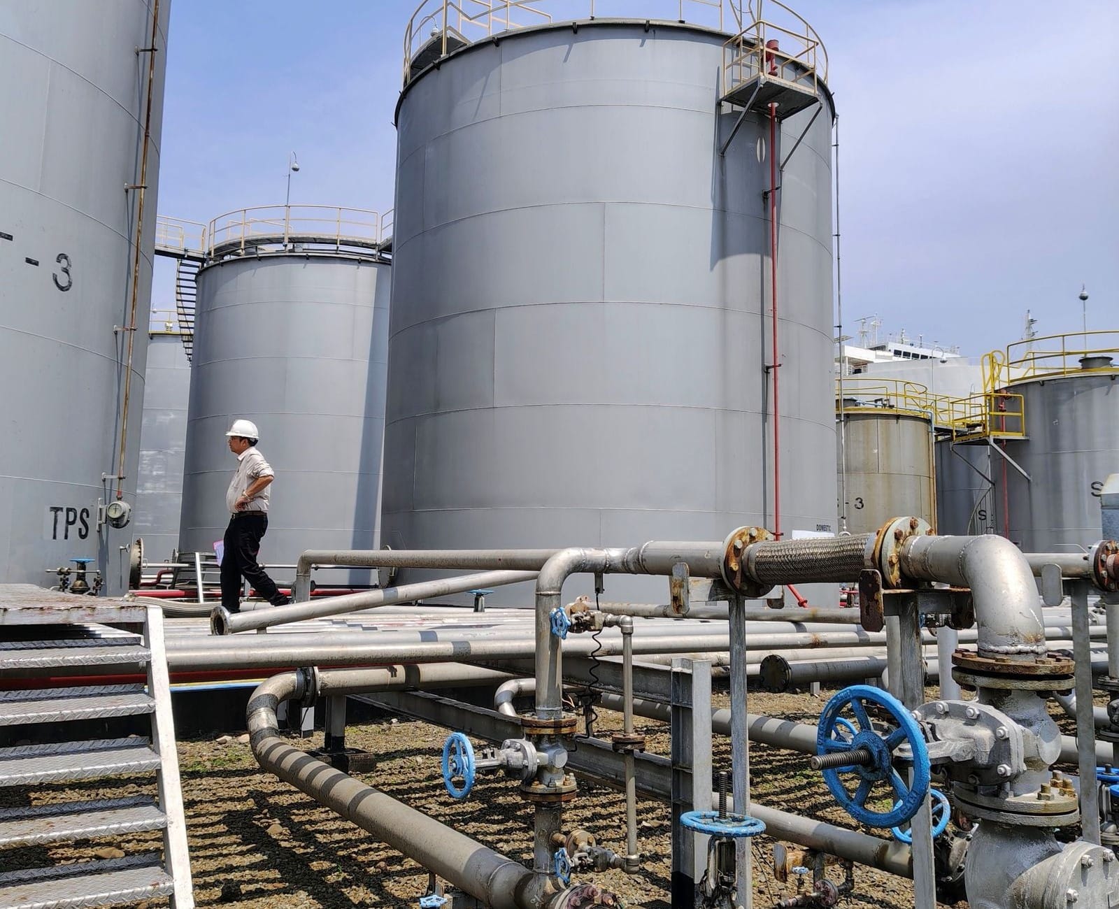 Worker walking past grey storage tanks and valved pipework on a gravel tank pad under a blue sky