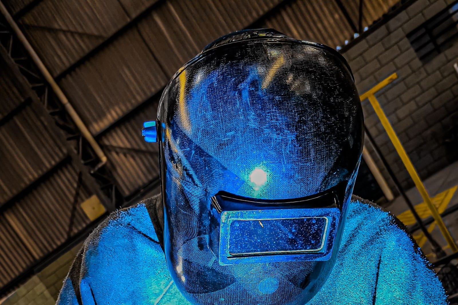 Welder in a full helmet and blue-lit jacket seen from above, with the arc reflected in the helmet visor inside a workshop