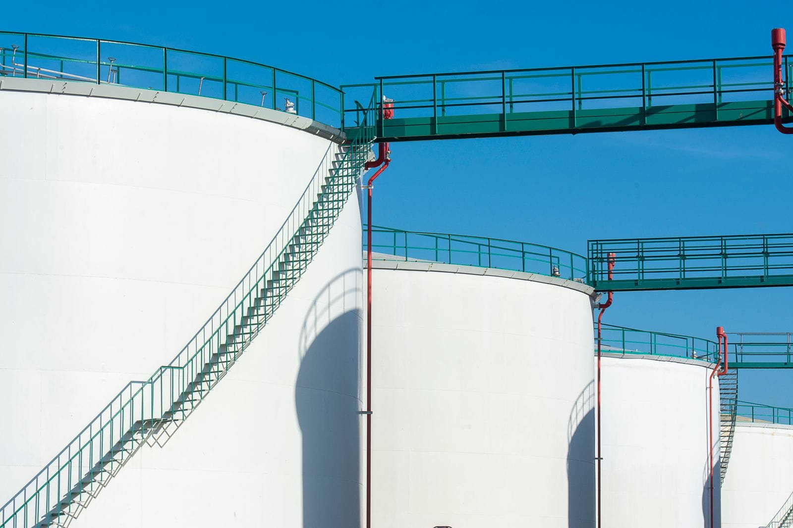 Large white storage tanks joined by a green elevated walkway and stairs against a clear blue sky